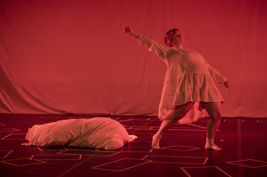 Eine Person tanzt auf der Bühne. Sie trägt ein weißes Kleid. Die Bühne ist rot beleuchtet. Auf dem Boden liegt ein weißes Kissen.